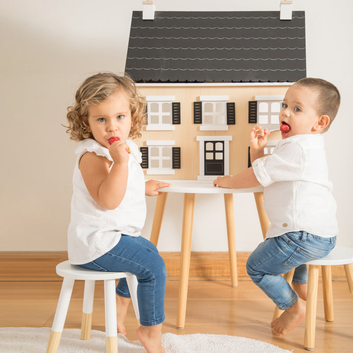 Dos niños jugando con una casa de muñecas grande y muebles infantiles (mesa y taburetes).