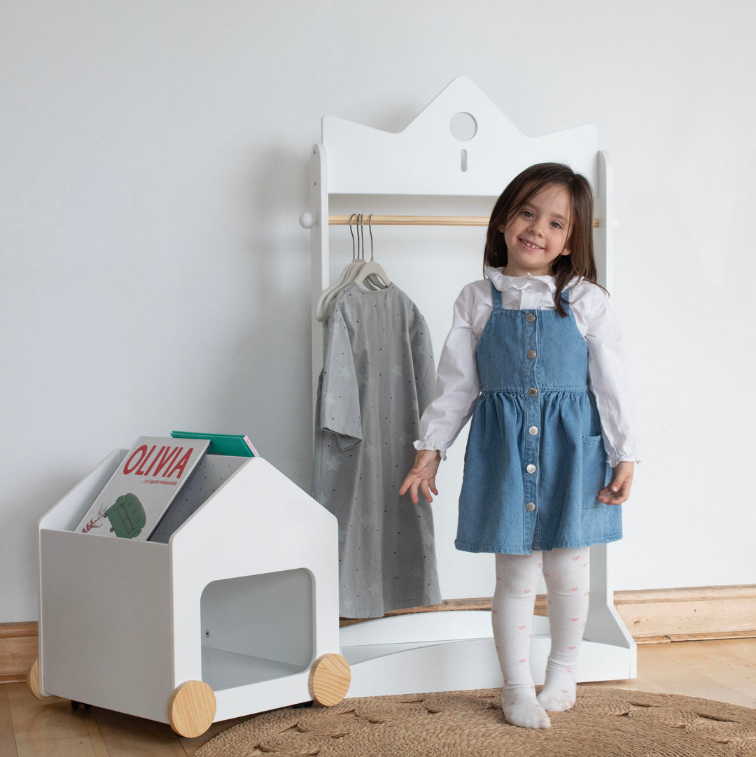 Niña junto a perchero infantil blanco y organizador de libros con ruedas, mobiliario Montessori ideal para dormitorios