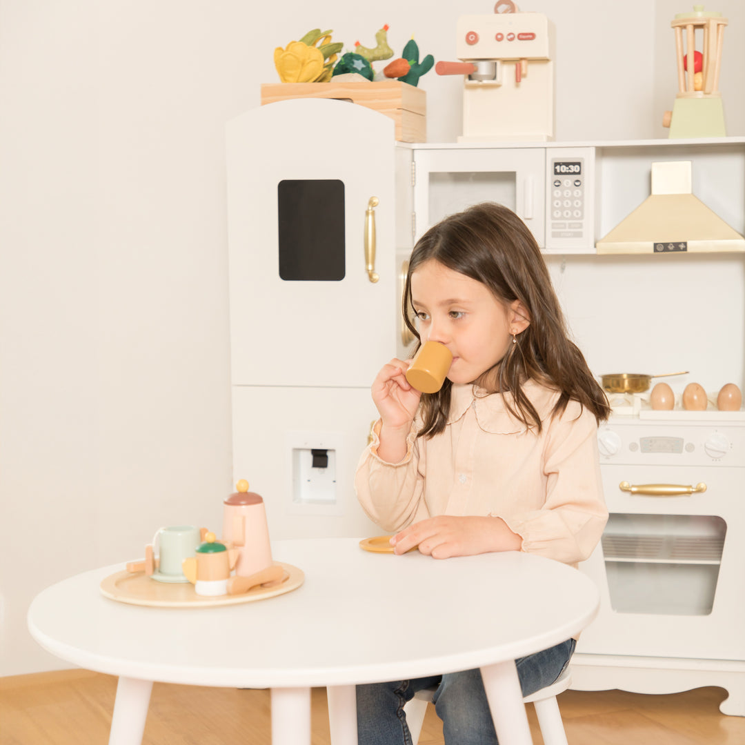 Niña jugando con set de té de madera en cocina de juguete blanca, fomentando el juego de roles y creatividad