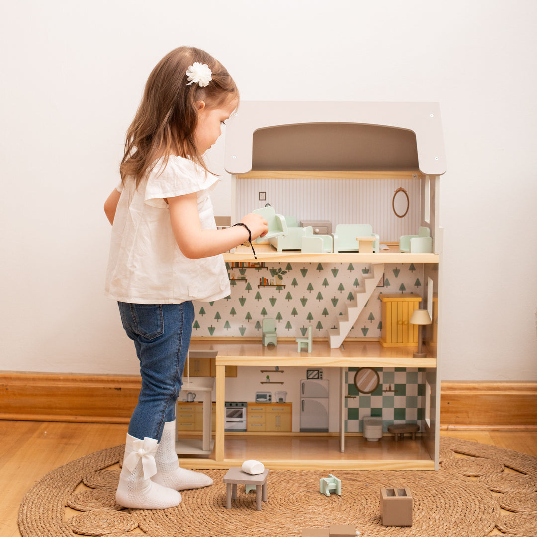 Niña jugando con la casa de muñecas de madera con tres pisos, techo blanco, muebles y figuras de madera.