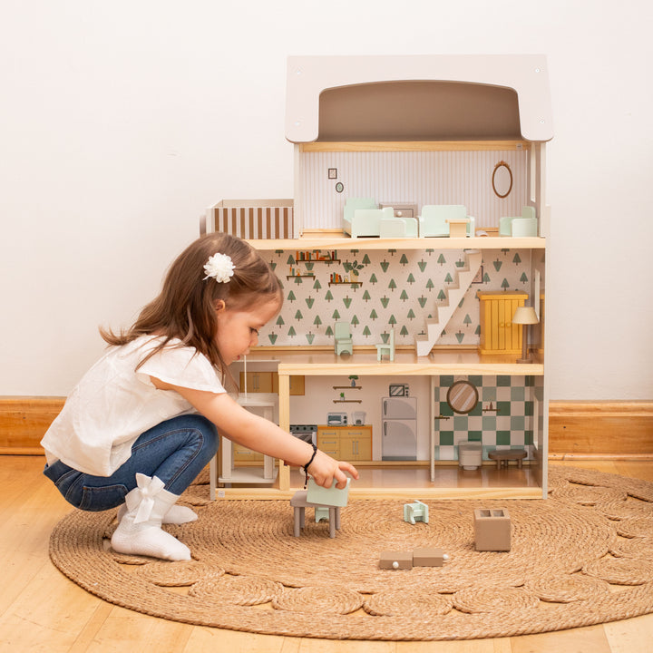 Niña jugando con la casa de muñecas de madera con tres pisos, techo blanco, muebles y figuras de madera.