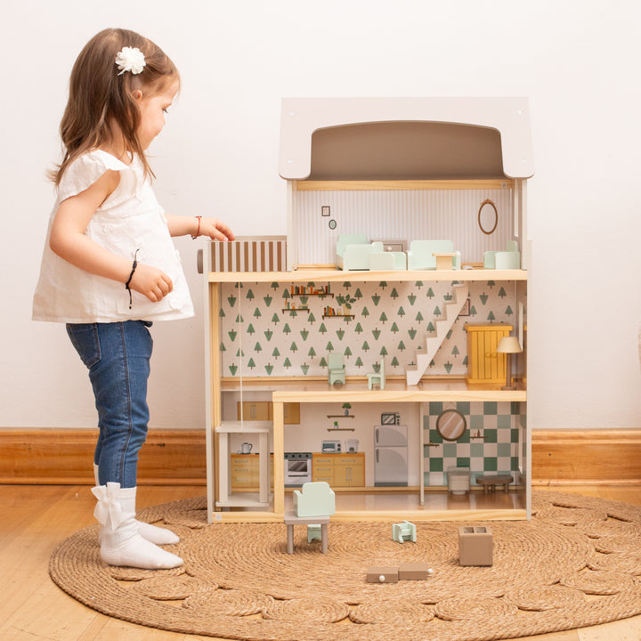 Niña jugando con la casa de muñecas de madera con tres pisos, techo blanco, muebles y figuras de madera.